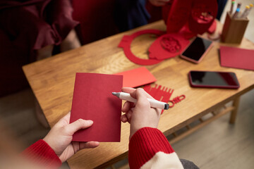 Fototapeta premium Asian young adult writing on red envelope with pen during Tet holiday, sitting at wooden table with other red envelopes, smartphones and festive decorations visible in background