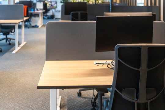 Contemporary office environment featuring wooden desks with monitors, ergonomic black office chairs, white privacy screens, and modern minimalist interior design with indirect lighting.