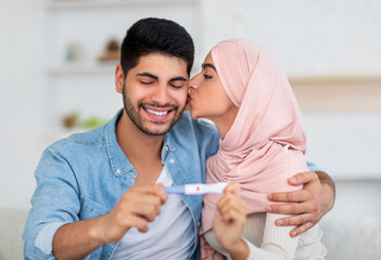 Happy arab woman kissing her husband with positive pregnancy test at home. Muslim couple happy to find out about their future baby, exchanging caresses indoors