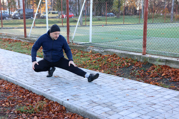 Man doing side leg stretch on a sidewalk near a sports field during an autumn workout