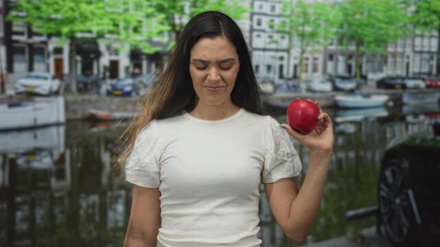 Woman cringes while holding red apple by amsterdam canal street with historic buildings visible; distaste.