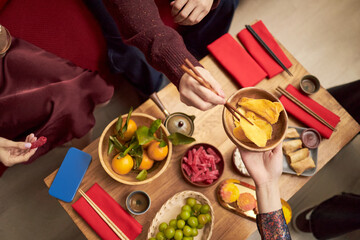 Fototapeta premium Asian young adults and teenagers sharing traditional holiday snacks using chopsticks, hands reaching over wooden table with dried fruit, citrus, sweets and festive red napkins during Tet celebration