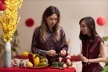 Two young adult Asian women preparing traditional offerings and lighting incense at decorated table...