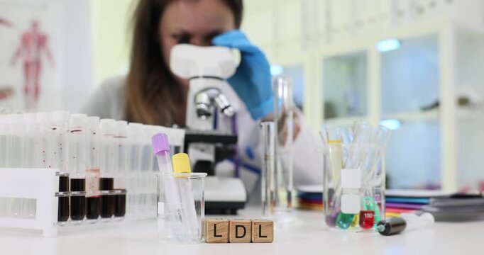 Woman laboratory worker examines LDL under modern microscope. Miniature wooden cubes on table clearly form abbreviation LDL near test tubes in rack