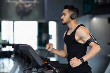 Fototapeta premium Muscular Arab Male Athlete Wearing Wireless Headphones Running On Treadmill At Gym, Sporty Millennial Middle Eastern Guy Listening Music During Workout, Enjoying Cardio Training, Copy Space