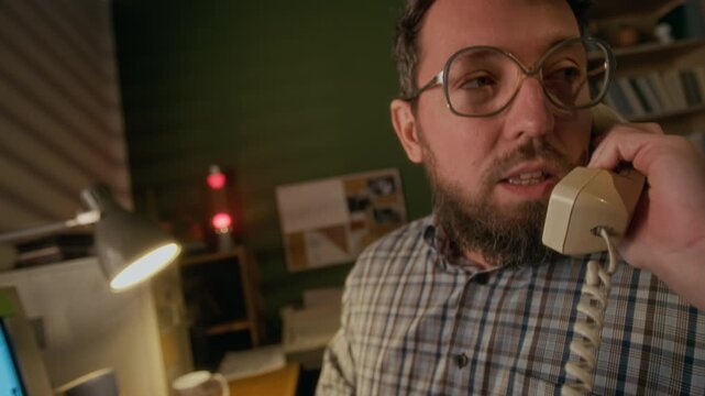 Medium shot of middle-aged Caucasian man working on vintage desktop computer at home, talking on landline telephone to colleague or manager, discussing business matters