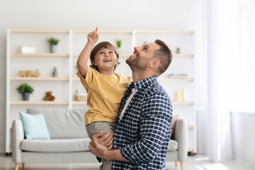 Fototapeta premium Daddy, look up. Excited little boy looking upwards with his father, child pointing with finger, man holding kid on hands, standing together at home