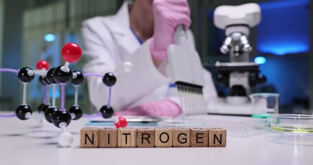 Word Nitrogen on wooden cubes by molecular structure model in chemical lab. Female assistant drips reagent into specimens at workplace with microscope - Powered by Adobe