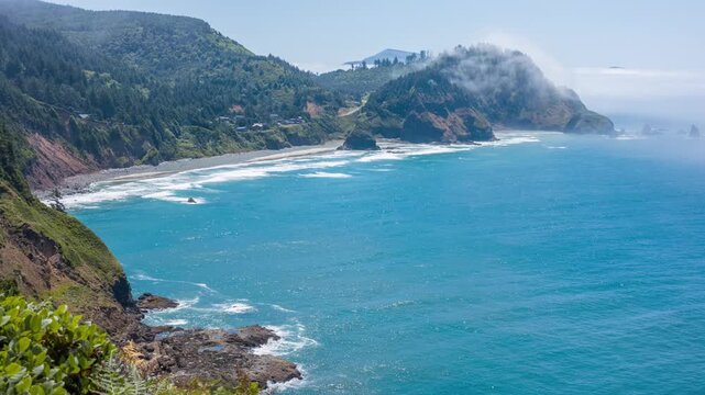 A scenic view of the rugged coastline and the Pacific Ocean from the Cape Meares State Scenic Viewpoint in Tillamook, Oregon. The image captures rolling hills covered in lush greenery, dramatic sea cl