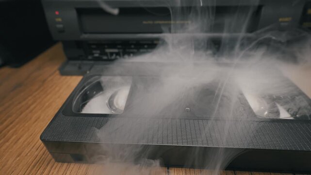 Black VHS cassette sits on wooden table emitting thick white smoke near blurry VCR player. Wispy steam rises from plastic housing indicating malfunction or overheating retro electronics.
