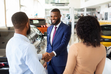 Fototapeta premium Car Dealership Concept. Salesperson Handshaking With Young Black Customers Couple In Showroom, African American Spouses Buying New Automobile In Modern Auto Salon, Selective Focus On Manager