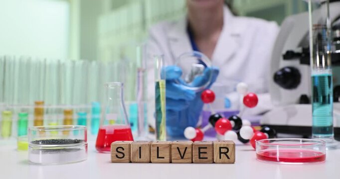 Woman laboratory worker holds Petri dish showing silver sample. Wooden cubes form Silver indicating substance currently under analysis by microscope