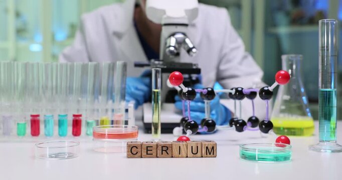 Laboratory worker studies prepared sample under high-precision microscope. Wooden cubes form word Cerium indicating analyzed element at test tubes