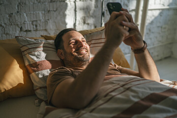 Smiling man waking up in bed checking smart phone