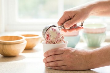 Person scoops ice cream into cup in a kitchen while bowls are placed on the counter and sunlight comes through the window