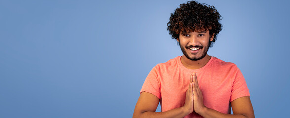 Man with curly hair stands with hands together in a greeting gesture. He smiles at the camera against a blue background. The setting is simple and bright, focusing on his welcoming expression.