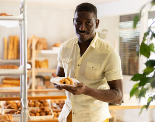 Positive African American man holding fresh tasty pastry, ready to purchase baked goods in bakery.