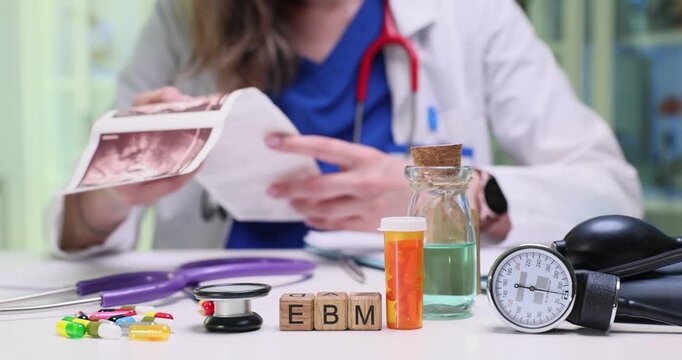 Doctor studies images noting subtle anatomical details. Cubes form abbreviation EBM highlighting evidence-based medicine approach used by physician