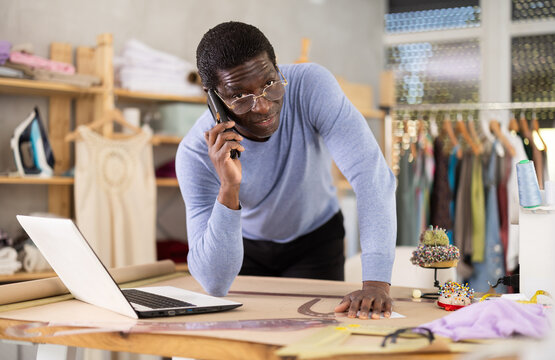African American male seamstress communicates with contractors using smartphone, orders fittings for sewing coat at laptop. Work issues, conference call to clarify issues