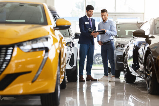 Handsome middle eastern guy standing among brand new cars, having conversation with cheerful sales assistant, looking at catalogue in dealer hands, checking auto features, full length