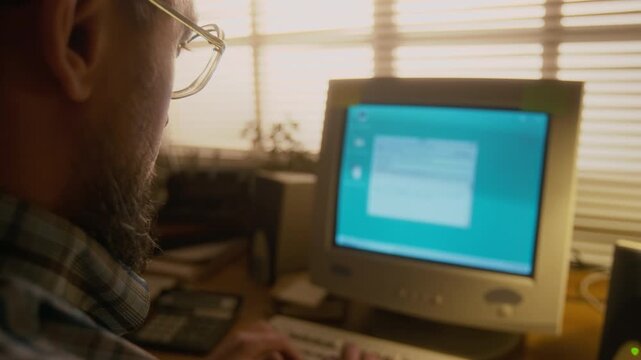 Over-shoulder shot of Caucasian man typing email on keyboard of retro desktop computer with flickering CRT monitor, calculator and documents on table, working at home by window covered with blinds