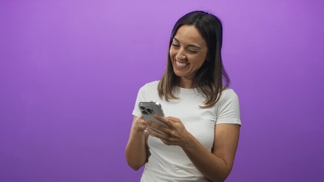 Woman tapping smartphone with finger in studio against purple backdrop, smiling while reading messages, wearing white t shirt; joy.