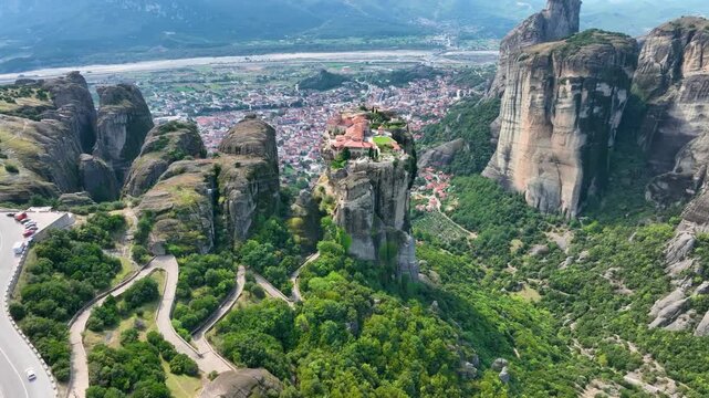 Wide aerial drone shot approaching the Holy Trinity Monastery in Meteora, Greece. Establishing shot revealing the winding roads, cliffs, and Kalabaka town in the valley below