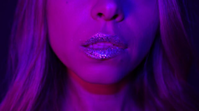 Close-up of young blonde woman biting glitter lips under pink and purple neon light. Sensual beauty moment with shimmering makeup, soft expression, glamour aesthetic and intimate mood