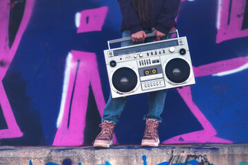 Young girl in streetwear jeans and boots carrying a retro boombox against graffiti wall, urban energy