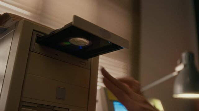 Close-up of 1990s Caucasian woman checking label on disk, then inserting into cd-rom drive on vintage computer, pressing button to close tray, while working on vintage desktop computer