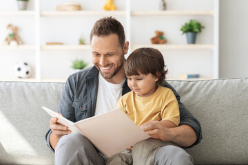 Naklejka premium Weekend activities with toddlers. Cheerful carying daddy reading book to little son, baby boy enjoying fairy tale on dad's knees, sitting on sofa at home
