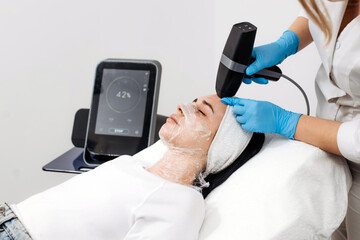 Female patient receiving RF lifting treatment at a beauty clinic, technician using a handheld device on her face with a digital display in the background showing treatment progress © wedmoments.stock