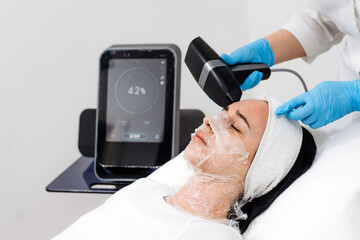 Woman receiving RF lifting treatment at a beauty clinic, technician using a handheld device on her face while monitoring progress on a digital display with copy space © wedmoments.stock