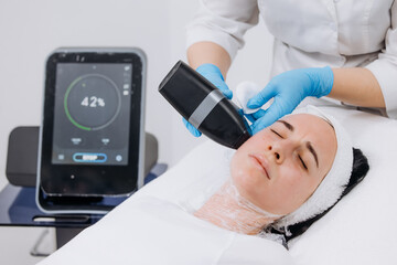 Female patient receiving RF lifting treatment at a beauty clinic, technician using a handheld device while a digital display shows treatment progress and settings with copy space © wedmoments.stock