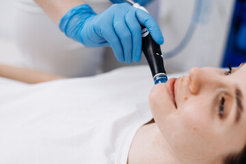 Fototapeta premium Cosmetologist performing hydrafacial treatment on a female client with blue gloves, close-up view of facial device and skin during skincare session in a clinical setting