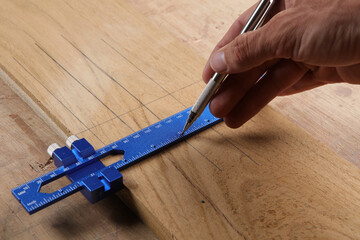 A hand uses a silver mechanical pencil and a blue precision gauge to mark a line across a wood...