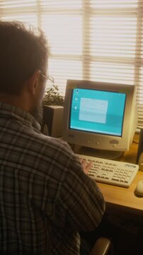 Vertical rear shot of businessman writing work email on vintage desktop computer with flickering blue screen, typing on keyboard, sitting at desk in front of window blinds, nostalgic 1990s vibe