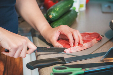 Photograph of a woman cooking in her kitchen; she is cutting meat with a sharp knife.
