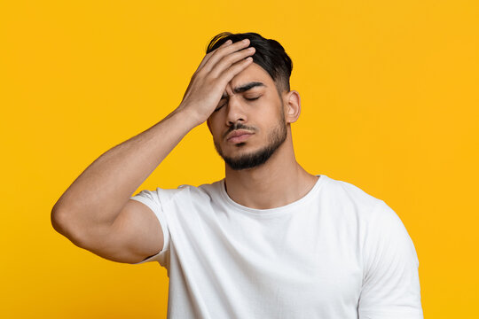 Exhausted bearded millennial arabic guy with closed eyes touching his head, feeling tired, yellow studio background, closeup shot. Human emotions and gestures, face expressions concept