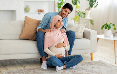 Obraz premium Pregnant muslim woman feeling relaxed, sitting on floor at living room and attentive husband massaging her shoulders, copy space. Full length portrait