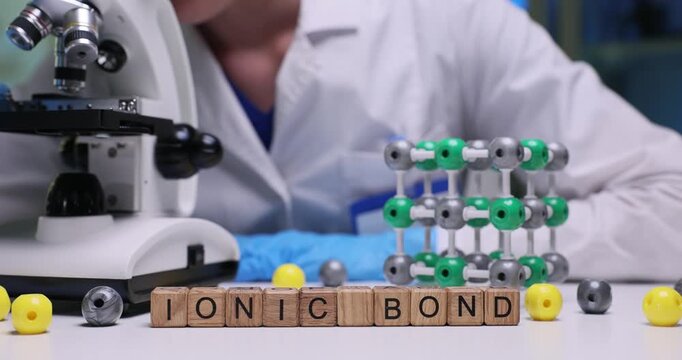 Text Ionic Bond on wood blocks with atoms and molecular structure model in lab. Female technician looks in microscope researching material at desk