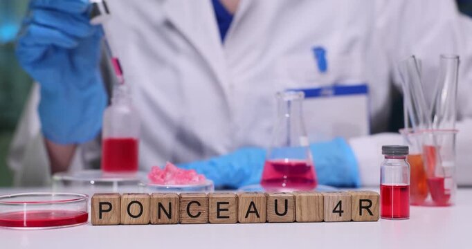 Text Ponceau 4R on wooden cubes and flasks with red liquid in chemical lab. Woman scientist uses pipette taking color fluid from bottle at desk