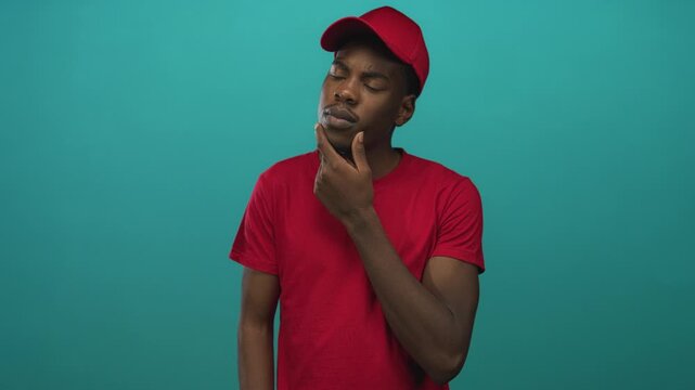 Man wearing red cap and t shirt with serious expression hand on chin in studio with teal background; introspection.