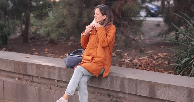Blonde woman buttons jacket neck to warm as sits on stone parapet slow motion. Female feels cold spending time in autumn park. Cold weather