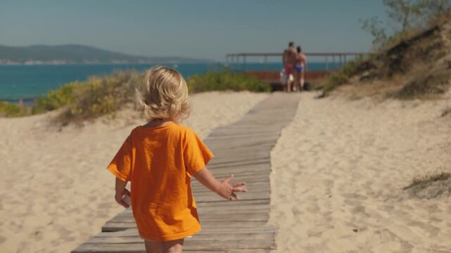 Child explores shoreline, Youngster strolls toward family, Small child on wooden path heading to family, Little kid in bright attire ventures along sands towards adults in sunshine