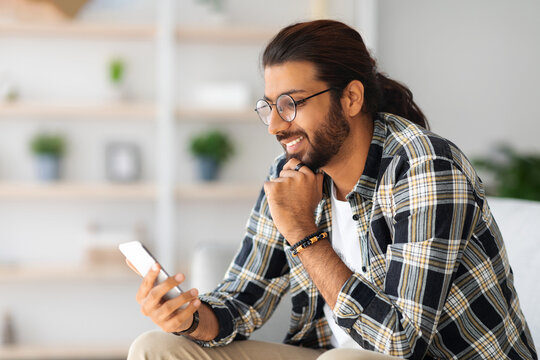 Cheerful long-haired middle-eastern guy with glasses sitting on couch, using smartphone, chatting with his girlfriend and smiling, happy millennial arab man chilling at home, side view, copy space