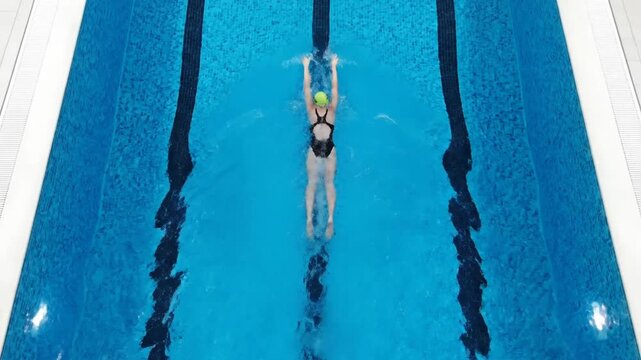 An overhead view captures a female athlete swimming the backstroke efficiently down a vibrant blue pool lane, showcasing the continuous arm movements over 8 seconds of footage.