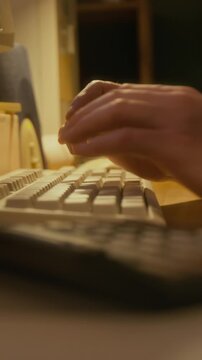 Vertical close-up of hands of anonymous man counting on calculator while working on report, typing on retro computer keyboard, in soft dim electric light in evening