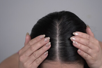 A close-up view shows a person using their hands with neutral-toned manicured nails to part their dark hair, highlighting the scalp and roots for examination or treatment.