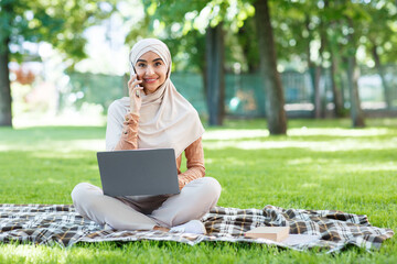 Workplace outdoor, modern business remotely, freelance, digital nomad. Cheerful young pretty arabian woman in hijab with laptop, speaks on phone, sits on plaid on green grass in park